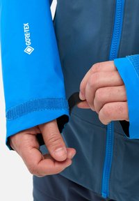 Die GORE-TEX-Jacke hat einen blauen Ärmel mit einem verstellbaren Klettverschluss am Bund und einen Reißverschluss vorne aus einem kontrastierenden dunklen Material.
