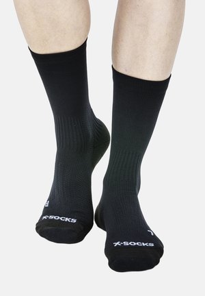 Persoon draagt zwarte sokken tot halverwege de kuit met het "X-SOCKS"-logo en aanwijzers voor rechter/ linker voet op een witte achtergrond.