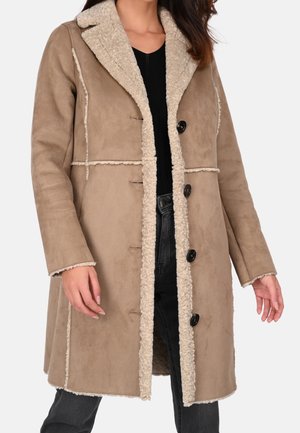 Femme portant un manteau beige en fausse peau lainée avec un grand col et des boutons noirs sur un haut noir et un jean foncé.