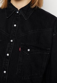 Musta farkkutakki, jossa on terävä kaulus, nepparikiinnitys ja yksi rintatasku punaisella tarralla. Teksturoitu kangas, kestävä muotoilu.