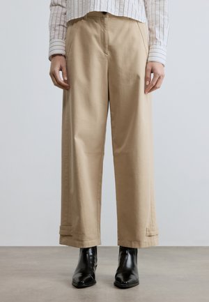 Person wearing beige wide-leg pants with cuff straps and black ankle boots on a gray floor and neutral background.