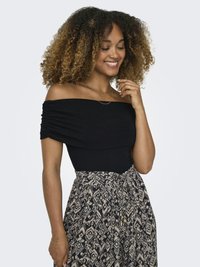 Usmievavá žena s kučeravými vlasmi, oblečená v čiernom topu odhalenom na ramenách a vysokopásovej sukni so vzorom, ruka blízko tváre, pozerá sa dole.