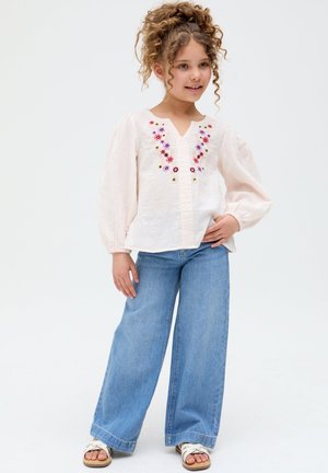 Young girl with curly hair wearing a light blouse with floral embroidery, wide-leg blue jeans, and white sandals, standing with hand on hip.