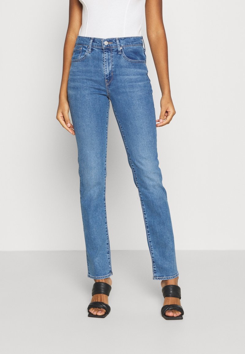 Femme portant un jean bleu slim et des sandales à talons noires ouvertes, debout devant un fond clair uni.