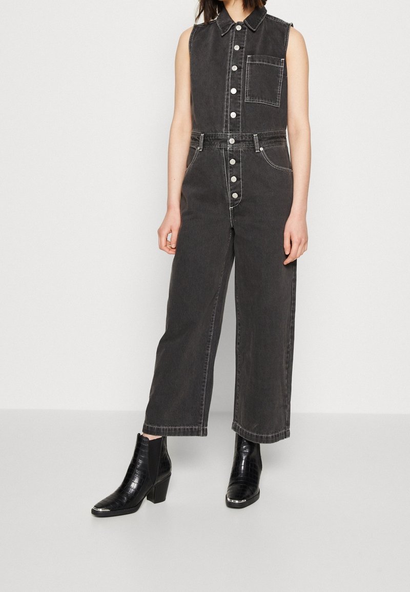 Femme portant une combinaison noire en denim sans manches avec des boutons blancs et des bottines noires à talons blocs, debout sur un fond uni.