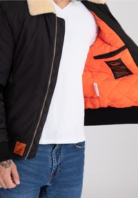 Svart bomberjacka med en fuskpälskrage, orange vadderad foder, dragkedjeslutning och etikett på vänster ärm. Bärs över en vit t-shirt.