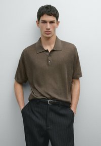 Hnědé polo tričko s krátkým rukávem a třemi knoflíky, spárované s černými kalhotami s proužky a tkaným opaskem, na neutrálním pozadí.