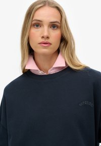 Sweatshirt marine à encolure ronde et logo brodé. Superposé sur une chemise rose à col. Texture lisse, design décontracté.