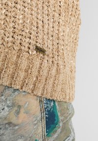 Beige pletený svetr s texturovaným vzorem a malou značkovou štítkou. Doplněn o vícebarevné kalhoty s abstraktními designovými prvky.