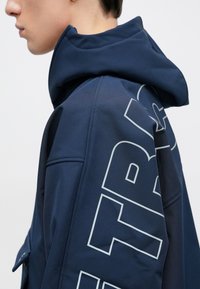 A tengerészkék kapucnis dzseki texturált mintával és nagy világosszürke felirattal az ujjon, sportos megjelenést kölcsönözve.