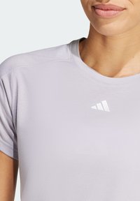 T-shirt de sport violet clair en tissu lisse, doté d'un col rond et d'un logo triangulaire blanc sur la poitrine.