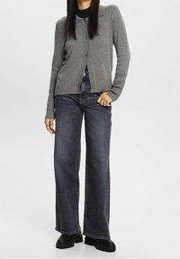 Šedý pletený cardigan s límcem, zapínáním na knoflíky vpředu a žebrovanými manžetami, v kombinaci s tmavými džínami širokého střihu a robustními černými botami.