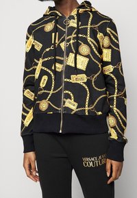 Sweat à capuche noir avec motif de chaînes dorées et d'étiquettes, doté d'un zip avant, de poignets côtelés et d'une taille côtelée ; associé à un pantalon noir.