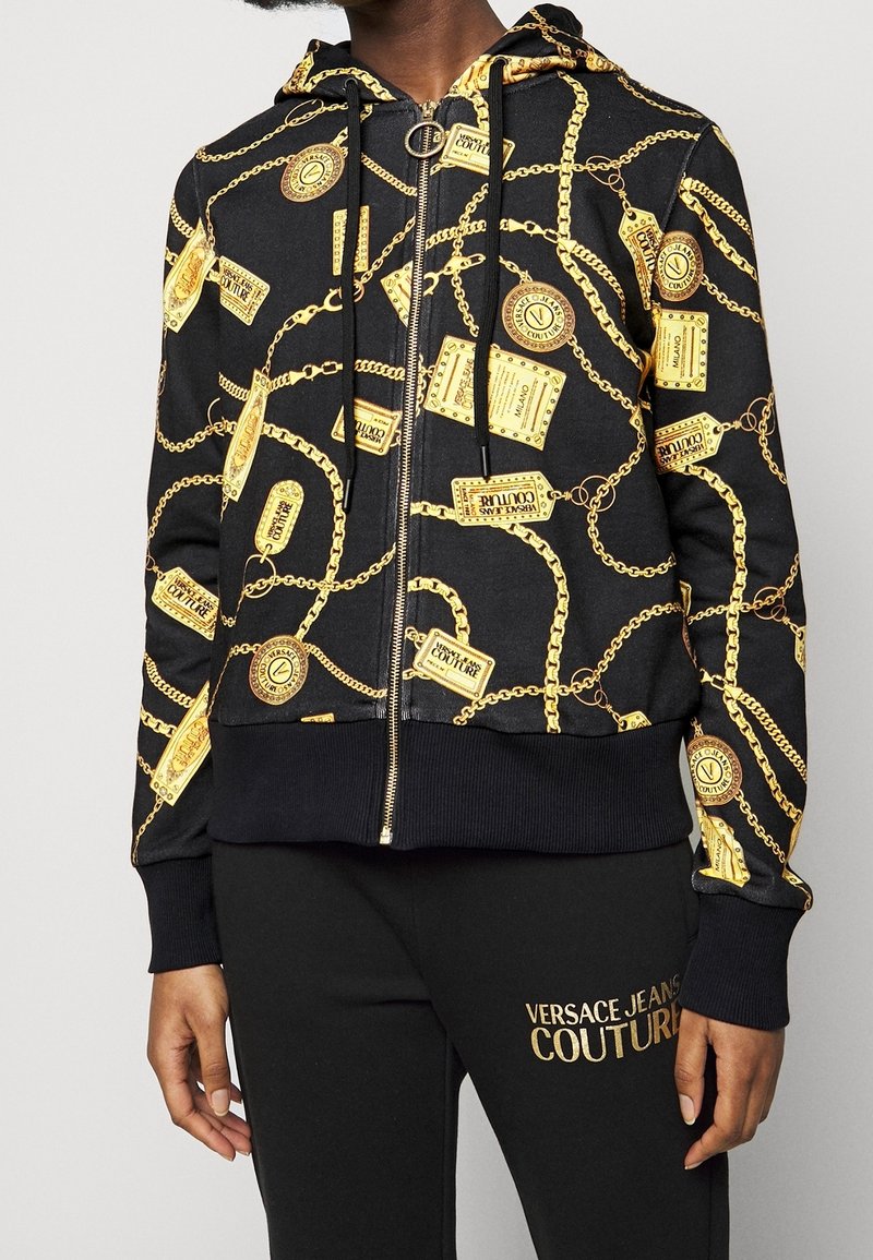 Sweat à capuche noir avec motif de chaînes dorées et d'étiquettes, doté d'un zip avant, de poignets côtelés et d'une taille côtelée ; associé à un pantalon noir.