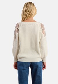 Maglione lavorato a maglia crema con maniche a sbuffo, caratterizzato da accenti di paillettes sulle spalle e orlo a coste. Abbinato a jeans in denim blu.