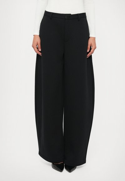 Pantalons larges noirs avec une texture lisse, taille haute et poches latérales, portés avec un haut ajusté à manches longues et des chaussures noires à talons pointus.