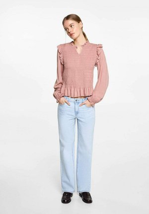 Staubrosa Smock-Top mit Rüschenschultern, V-Ausschnitt und ausgestelltem Saum. Kombiniert mit hellblauen gerade geschnittenen Jeans und schwarzen Schuhen.