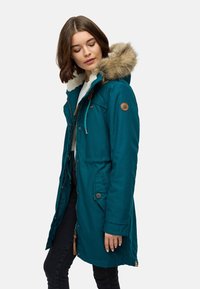 Parka teal con cappuccio bordato in pelliccia, chiusura con zip e bottoni, due tasche laterali e patch con logo sul petto. Tessuto testurizzato con finitura liscia.