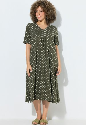 Kobieta z kręconymi włosami ubrana w oliwkową sukienkę midi z białymi wzorami gwiazd oraz oliwkowe plecione sandały wsuwane, stojąca przy białej ścianie.