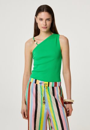 Woman wearing a green one-shoulder top with beaded strap and colorful vertical striped pants, standing against a plain background.