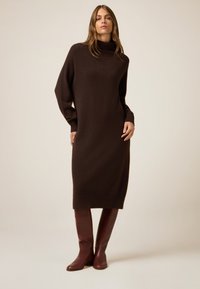 Un vestido de punto largo y marrón oscuro con un alto cuello cisne, mangas de gran tamaño y dobladillo acanalado; combinado con botas de cuero burdeos hasta la rodilla.