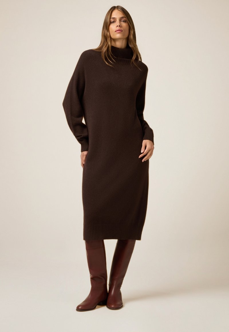Un vestido de punto largo y marrón oscuro con un alto cuello cisne, mangas de gran tamaño y dobladillo acanalado; combinado con botas de cuero burdeos hasta la rodilla.