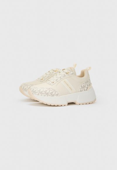 Zapatillas deportivas beige con una parte superior texturizada, herrajes dorados y un patrón de logotipo repetido, con una suela blanca gruesa y un tirador.