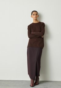 Braunes Langarmoberteil, kombiniert mit einem dunkelvioletten, gerippten Rock mit Schlitzen an der Seite, getragen mit braunen Stiefeletten. Glatte Textur.