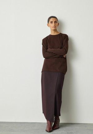 Braunes Langarmoberteil, kombiniert mit einem dunkelvioletten, gerippten Rock mit Schlitzen an der Seite, getragen mit braunen Stiefeletten. Glatte Textur.