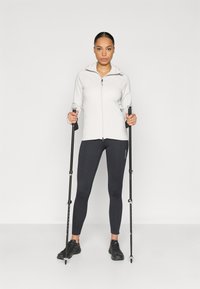 Hellgrauer Kapuzenpullover mit Reißverschluss, kombiniert mit schwarzen Leggings und schwarzen Schuhen. Die Person hält zwei Trekkingstöcke.