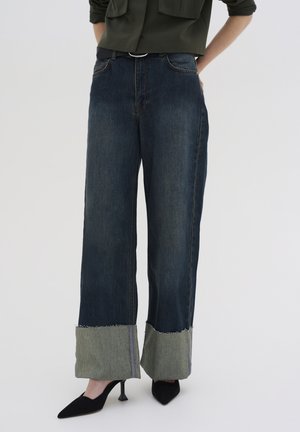 Široke denim hlače v temno modri barvi, z naravnim robom v zeleni barvi na koncu. Posebno šivanje dodaja teksturo. V kombinaciji s črnimi petami.