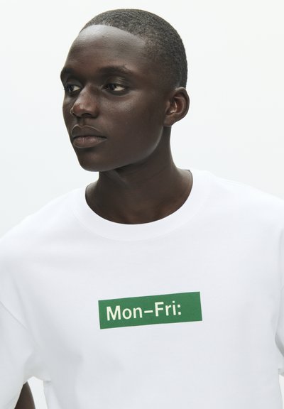 Hvit bomulls-T-skjorte med grønn rektangulær tekst som viser "Man-Fre:" i midten. Rund hals og korte ermer.