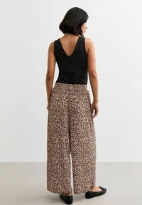 Schwarzes, strukturiertes, ärmelloses Oberteil und weit geschnittene Hose mit beigem Hintergrund und dunklem Leopardenmuster. Schwarze flache Schuhe runden das Outfit ab.
