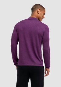 Man die glimlacht, met zijn rug naar de camera, in een paarse lange mouwtrui en zwarte broek, tegen een effen lichte achtergrond.