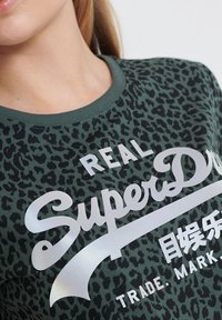 Zielona koszulka w grochy z nadrukiem lamparta, z okrągłym dekoltem i krótkimi rękawami. Posiada srebrny napis "REAL SuperDry" oraz gładką fakturę.