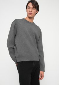 Grå genser laget av mykt strikket stoff med rund hals og lange ermer. Litt løs passform, parret med svarte jeans. Enkelt design.