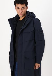 Parka bleu marine avec capuche, plusieurs poches à l'avant et une poche zippée sur le bras. Texture lisse et matériau durable.