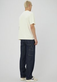 T-shirt crème à manches courtes associé à un jean large bleu foncé avec des coutures jaunes contrastantes et des poches latérales. Des baskets complètent le look.