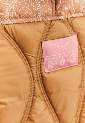 Brązowa pikowana tkanina z różową wszywką z zamkniętym okiem i tekstem, umieszczona wewnątrz podszewki kurtki lub torby.