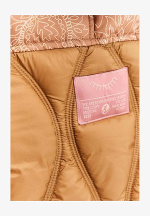 Brązowa pikowana tkanina z różową wszywką z zamkniętym okiem i tekstem, umieszczona wewnątrz podszewki kurtki lub torby.