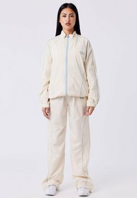 Lichtbeige trainingspak gemaakt van glad, lichtgewicht materiaal. Kenmerken zijn blauwe accentstiksels, een hoge kraag en elastische manchetten aan de mouwen en broek.