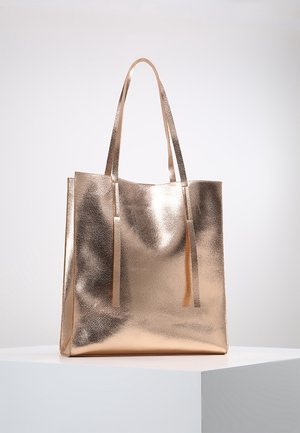 Duża metaliczna torba typu tote w kolorze różowego złota z dwoma długimi uchwytami, wystawiona na białym stojaku na jednolitym tle.