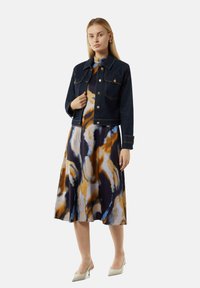 Denimjacke mit Knopfverschluss, zwei Brusttaschen; darunter ein fließendes Kleid mit einem blauen, orangefarbenen und cremefarbenen abstrakten Muster.