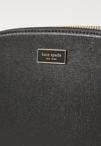 Portafoglio in pelle nera con texture, dotato di una placca con logo "kate spade" in tono oro. Chiusura a zip e motivo in rilievo sulla superficie.