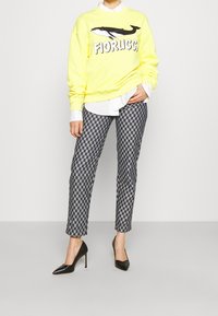 Sudadera amarilla con gráfico de "Fiorucci" y ballena, camisa blanca debajo, pantalones de cuadros grises y azules, tacones altos negros.