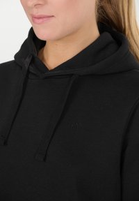 Lähikuva musta huppari päällä olevan henkilön, jossa on nyörit ja rintapielessä hienovarainen brodeerattu logo.