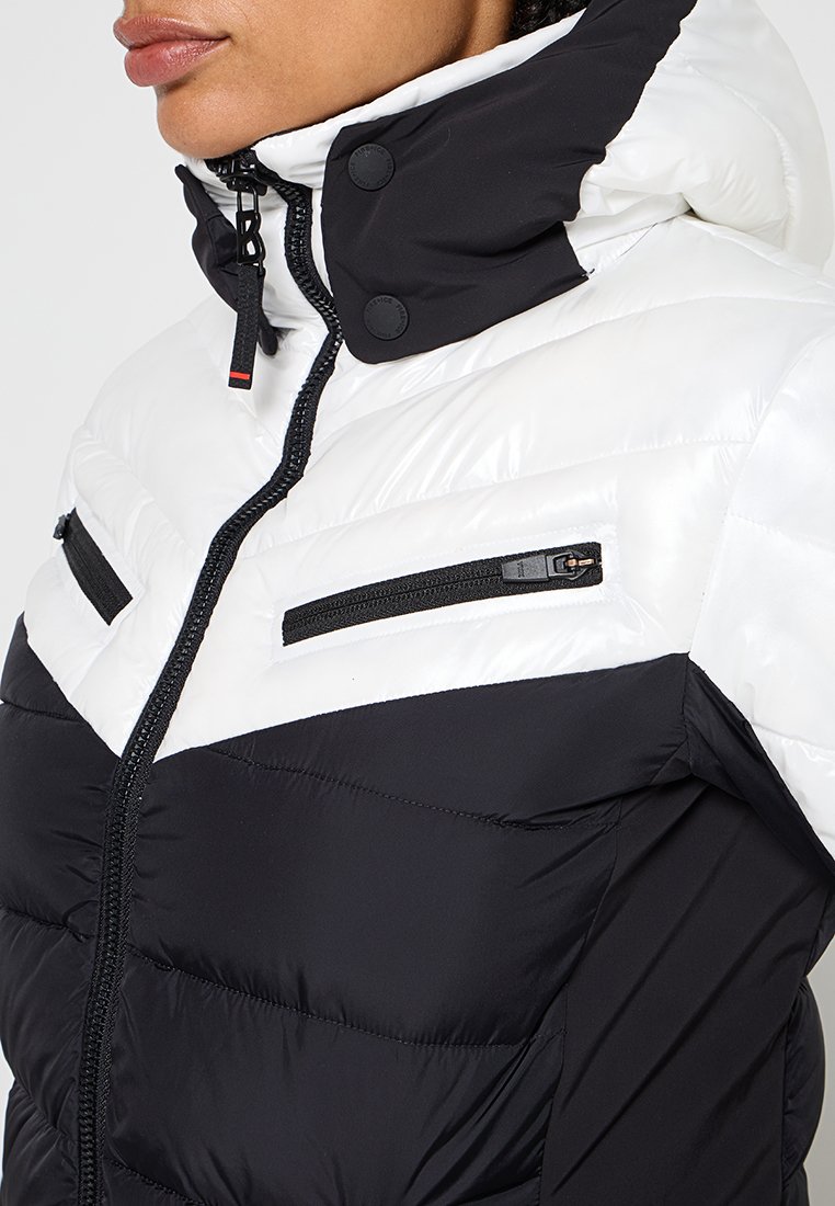 Padded Jacke mit einem Schwarz-Weiß-Farbschema, Reißverschlusstaschen und einem hohen Kragen mit Knopfakzenten. Hergestellt aus leichtem Material.