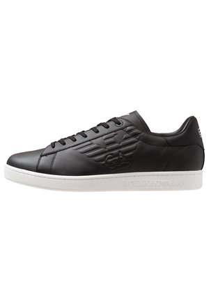Baskets basses noires avec lacets noirs, logo embossé sur le côté et semelle blanche avec le nom de la marque embossé sur le bord.