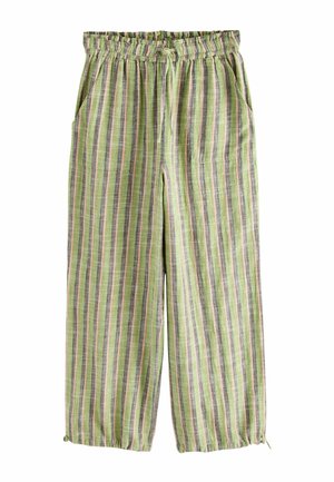 Žena v žltom halter top, zelených pruhovaných nohaviciach, slamák, a sandále kráča po zakrivenej kamennej ceste pri mori.