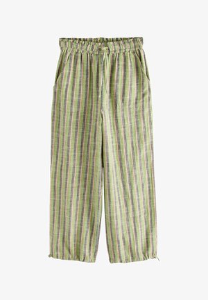 Kobieta w żółtym topie na szyję, zielonych spodniach w paski, słomkowym kapeluszu i sandałach, idąca wzdłuż krętej kamiennej ścieżki nad morzem.